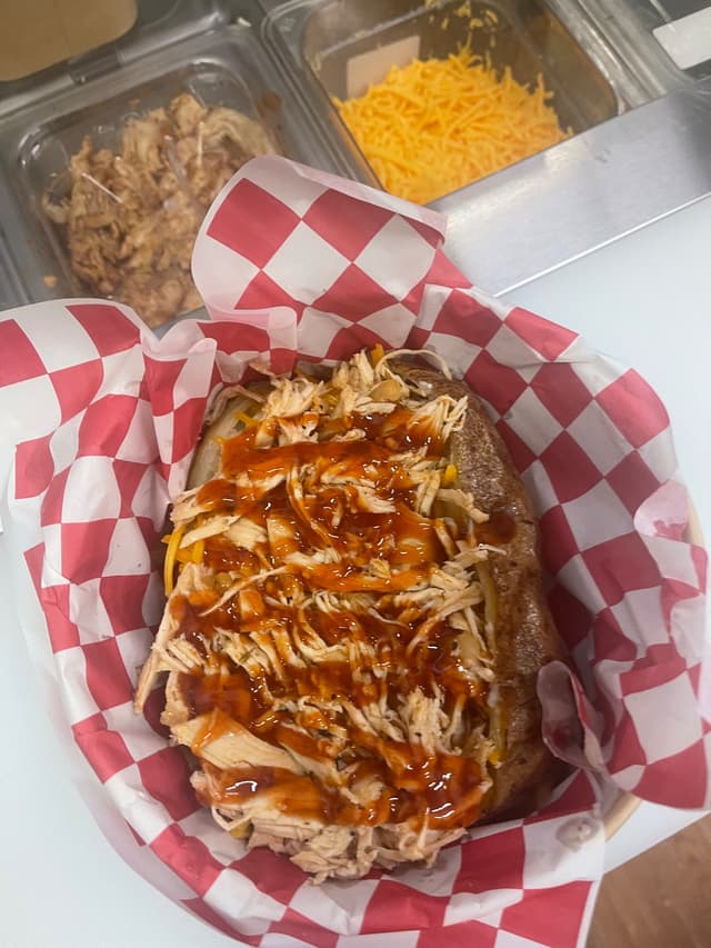 Loaded baked potato with shredded BBQ chicken, cheese, and sauce in red checkered paper